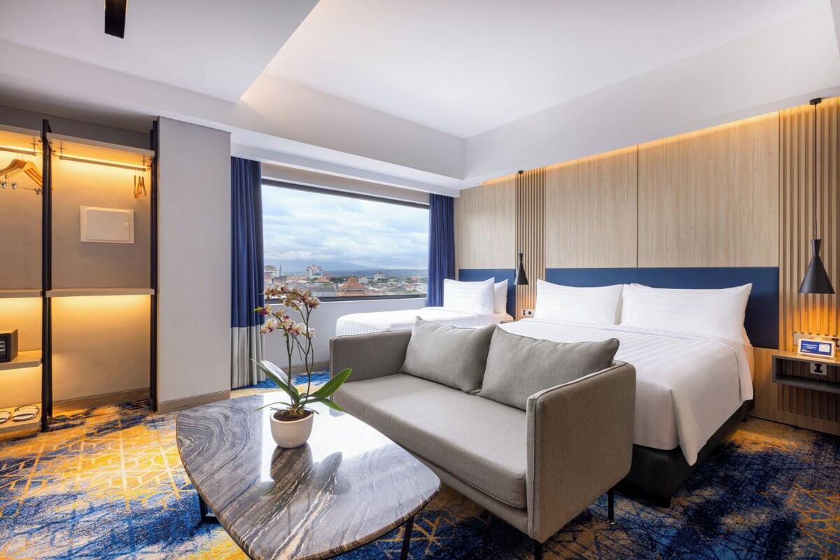 A hotel room with a bed, a sofa, a small table, a potted plant, and a view of the city.