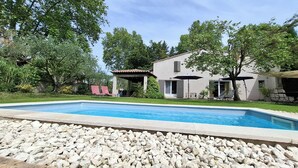 En swimmingpool omgivet af hvide sten, et toetagers hus med terrasse, og grønne træer.