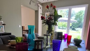 Et spiseområde med et bord dækket til to personer, en vase med blomster og et vindue med udsigt til grøn vegetation.