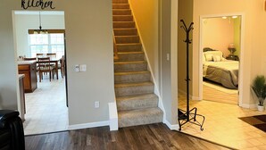 Un escalier mène à une chambre à coucher qui contient un lit, une table de chevet et une lampe. Un porte-manteau se trouve près de l’escalier.