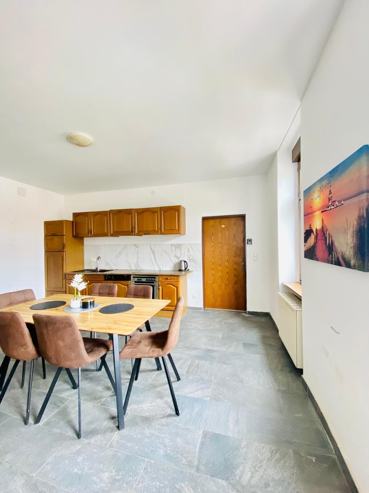 A modern kitchen with wooden cabinets, a dining area with a wooden table and chairs, and a painting on the wall.