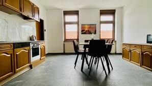 Une cuisine spacieuse avec des meubles de rangement en bois, une table à manger avec des chaises et deux fenêtres équipées de stores.