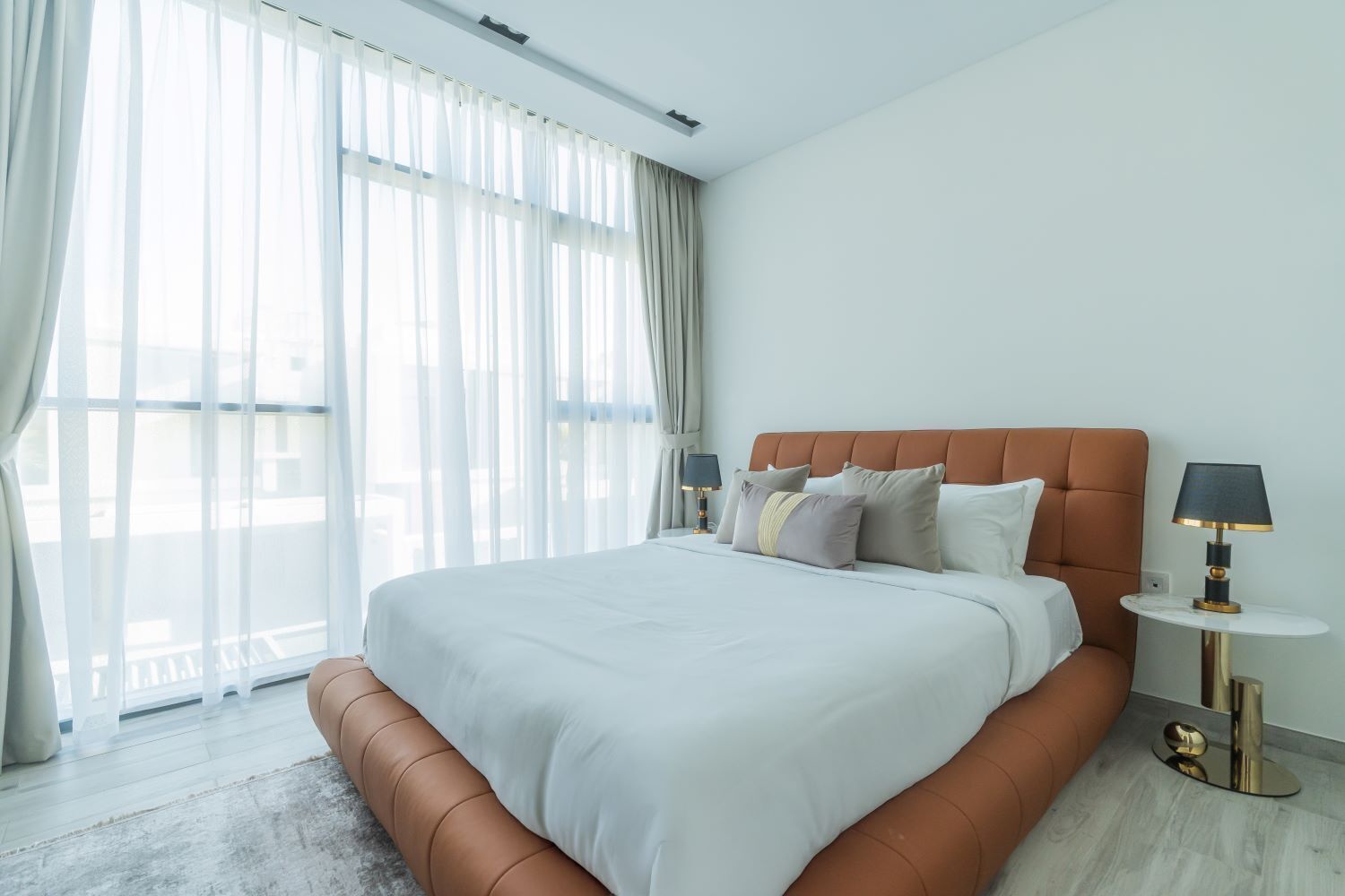 A modern hotel room with a large bed, a round side table, and a view of the city through sheer curtains.