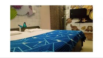 A hotel room with a bed covered in a blue patterned bedspread, a white pillow, and a blue pillow. There is a wooden headboard, a wall-mounted TV, and decorative wall art.