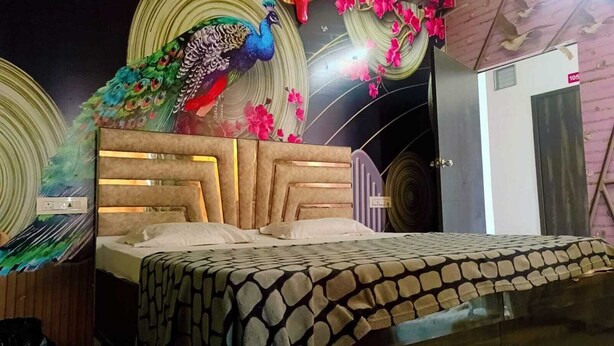 A bed with a patterned bedspread, a wooden headboard with light fixtures, and decorative wall art including a peacock and floral designs. - MS GRAND HOTEL (Bhiwani)