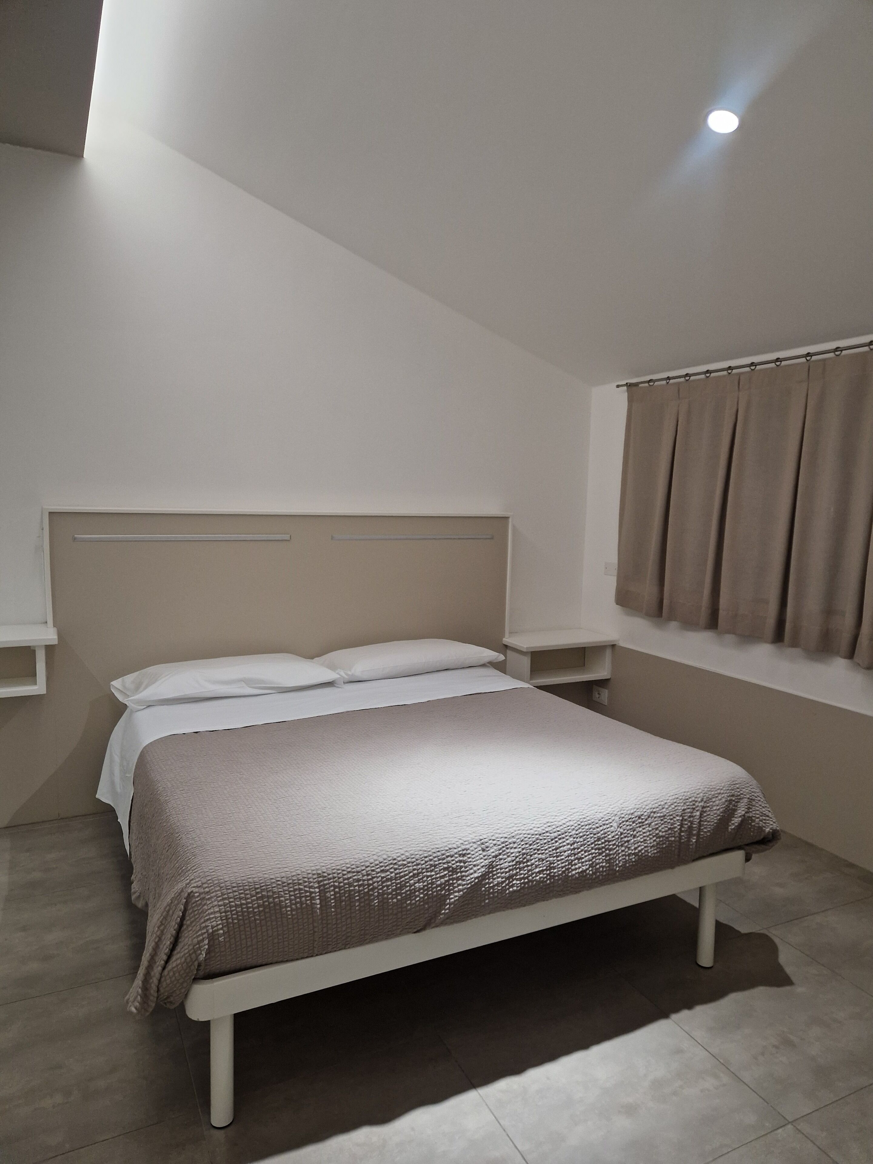 A bedroom with a bed, a headboard, a nightstand, a window with curtains, and a ceiling light.