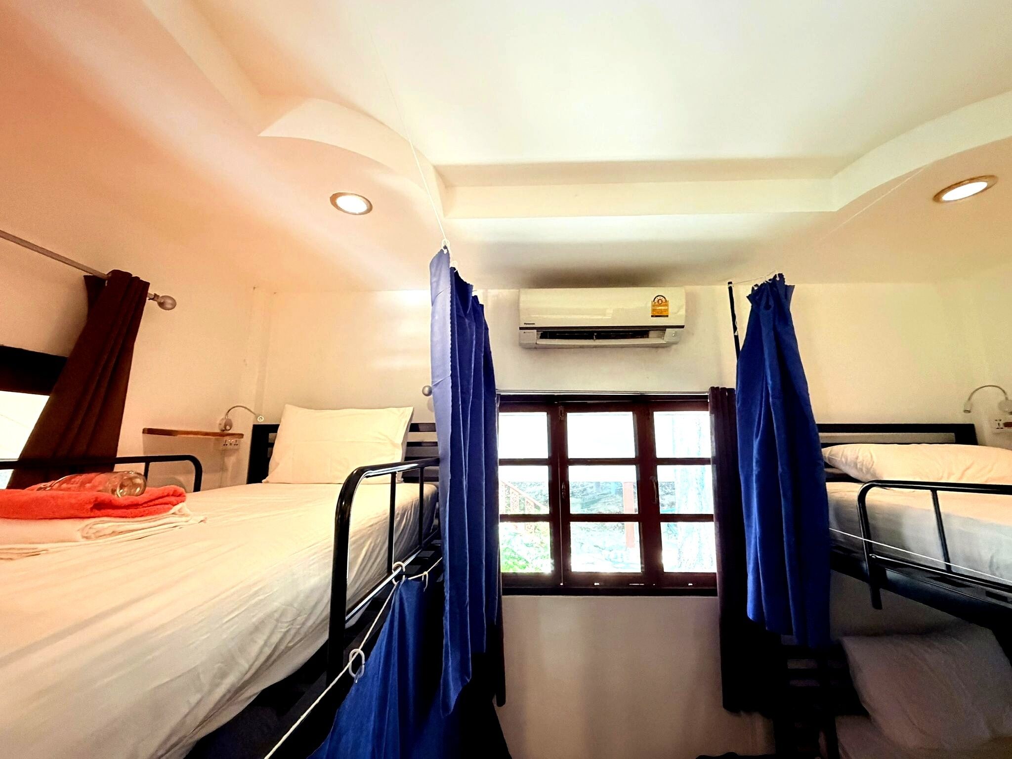 A hotel room with two beds, a window with curtains, an air conditioning unit, and a view of the outside.