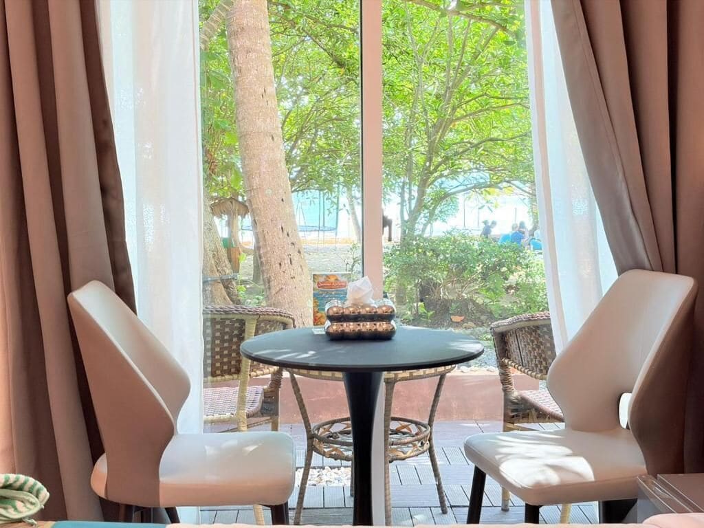 A room with a round table and two chairs, a window with curtains, and a view of greenery outside.