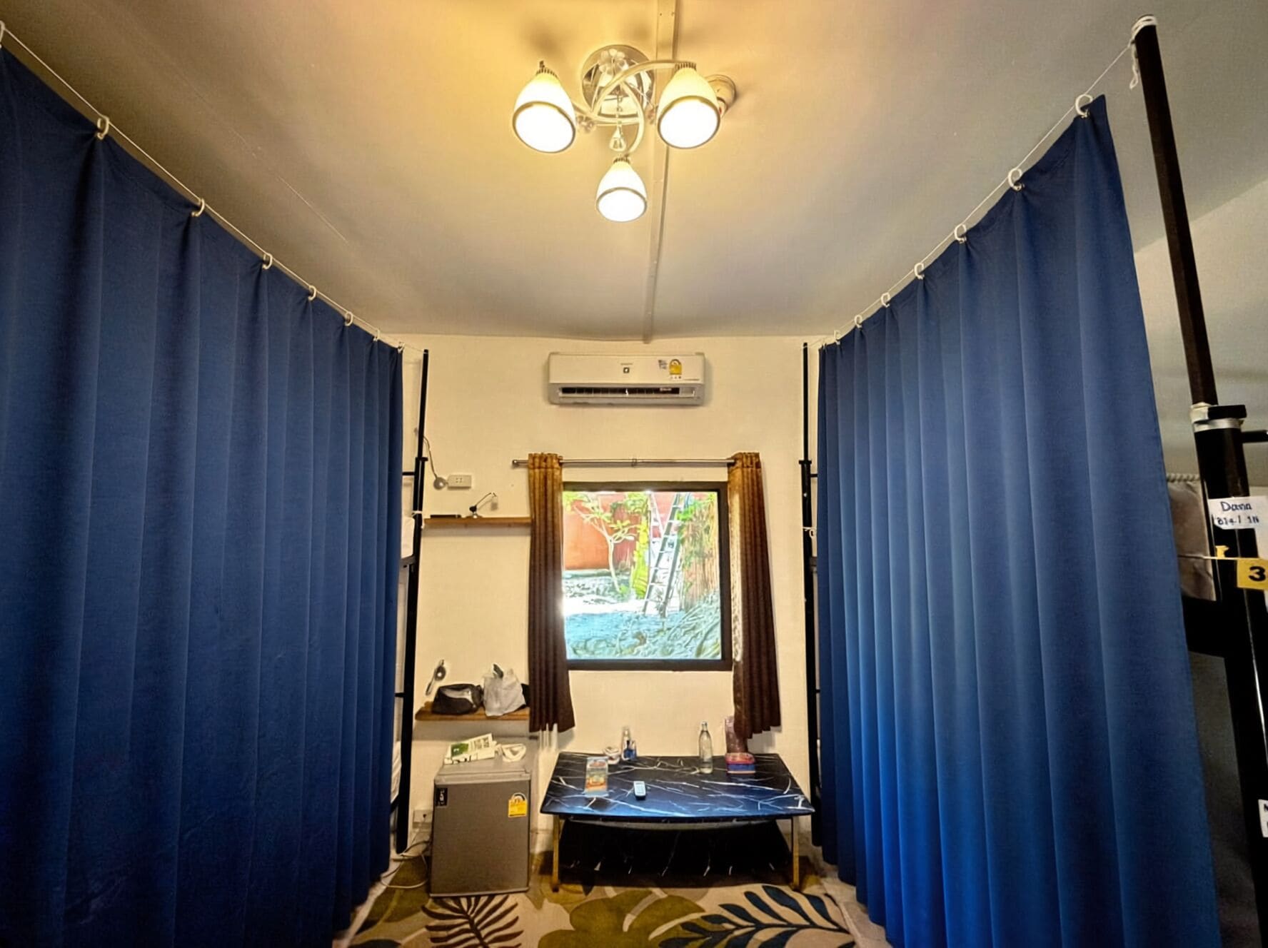 A room with a bed, a small table, a desk, an air conditioning unit, and curtains.