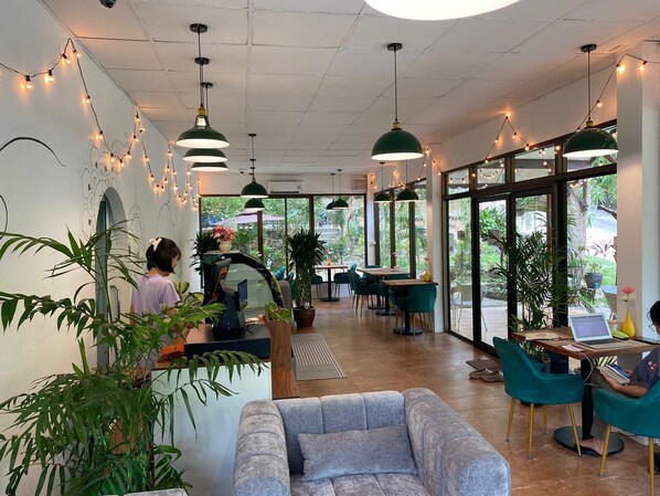 A modern cafe with green pendant lights, wooden tables, and chairs. There are potted plants and a sofa.