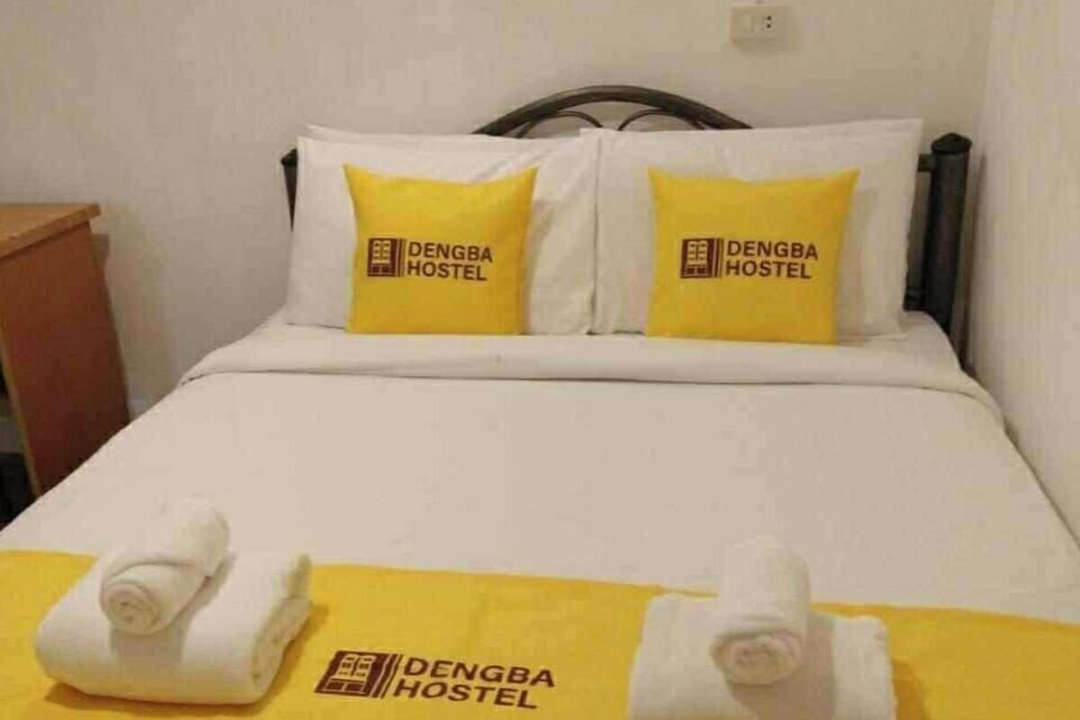 A neatly made bed with two pillows and a yellow towel on each side, featuring the logo of Dengba Hostel.