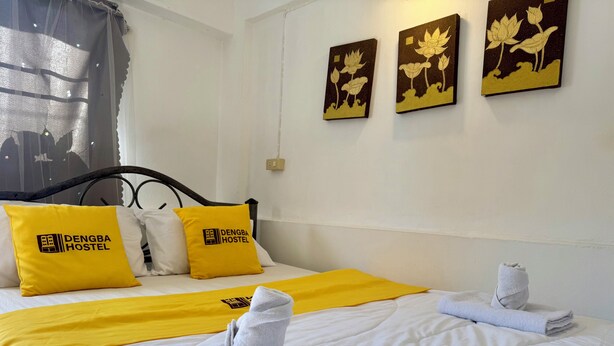 Una cama bien hecha con una colcha amarilla, dos almohadas con el logotipo 'DENGBA HOSTEL' y dos toallas dobladas sobre la cama.