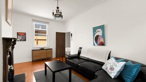 A living room with a black leather sofa, a wooden coffee table, a chandelier, and a painting of a bird on the wall. - Art Deco Era Guest Cottage with Off Street Parking (Northcote)