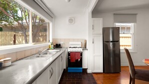 A kitchen with a stainless steel sink, a stove, a refrigerator, and a window overlooking a backyard. - Art Deco Era Guest Cottage with Off Street Parking (Northcote)