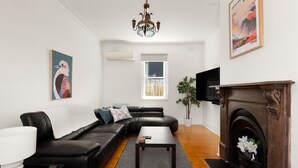 A modern living room with a black leather sofa, a small table, a fireplace, and a large painting on the wall. - Art Deco Era Guest Cottage with Off Street Parking (Northcote)
