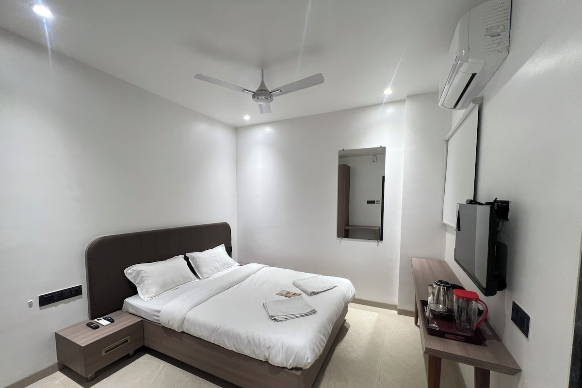 A modern hotel room with a bed, bedside table, nightstand, ceiling fan, air conditioning unit, TV, and a small table with a coffee maker.