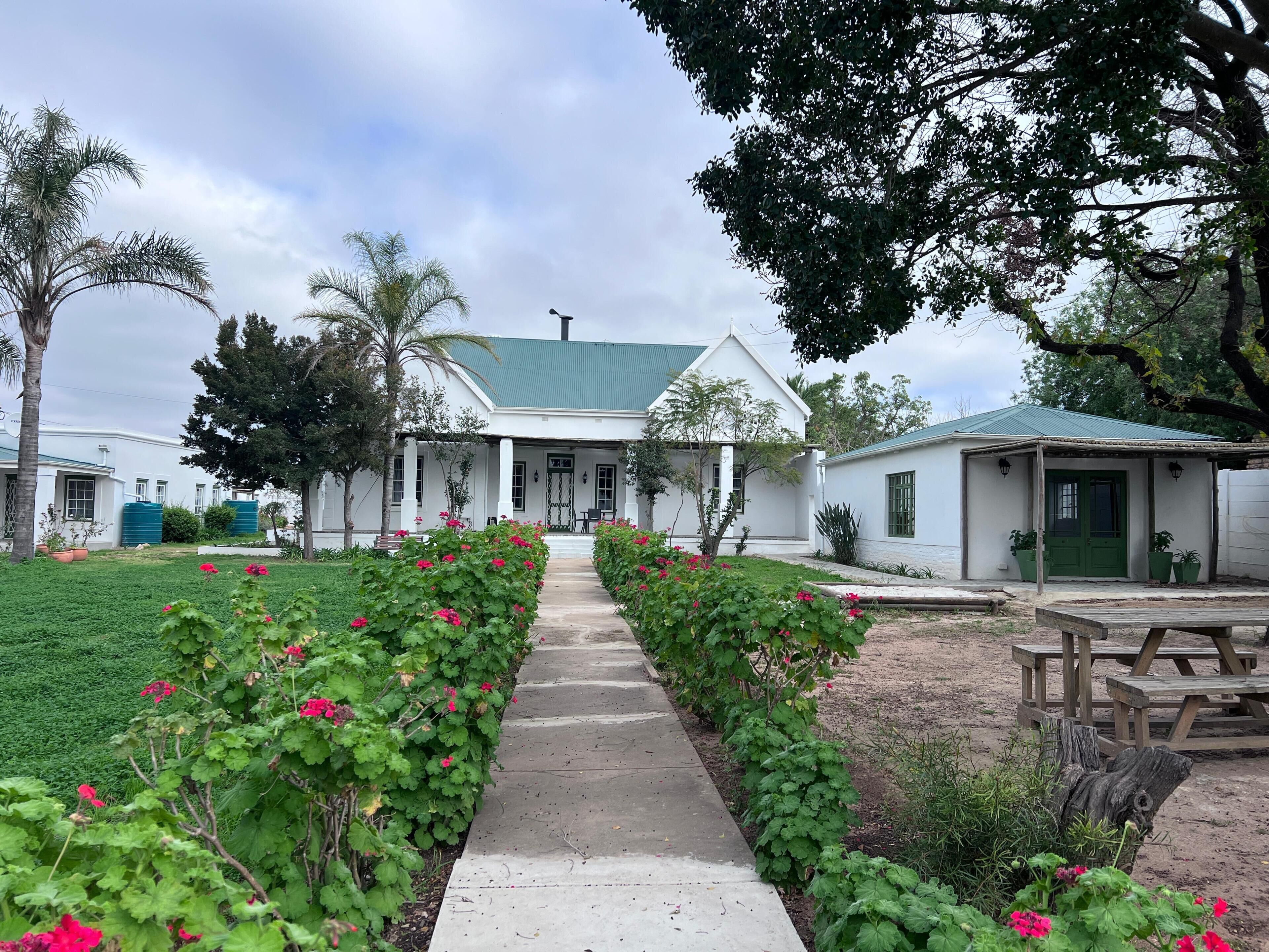 Un bâtiment blanc surmonté d’un toit végétalisé, avec un chemin pavé et un jardin où poussent des fleurs roses.