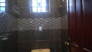 A bathroom with a toilet, a shower, and a window with a decorative grille. - Premier Suites Isiolo (Isiolo)