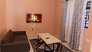 A living room with a sofa, a table, a TV mounted on the wall, and a window with curtains. - Premier Suites Isiolo (Isiolo)