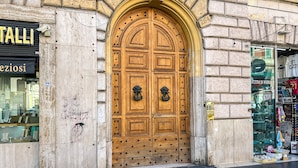 Une grande porte en bois surmontée d’une arche décorative, un magasin aux vitrines en verre et un vendeur ambulant présentant divers articles à l’extérieur.