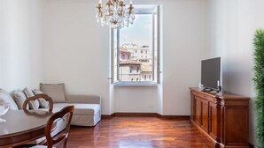 Une pièce comprenant une table à manger, des chaises, un canapé, un téléviseur posé sur un meuble en bois et une grande fenêtre donnant sur des immeubles.