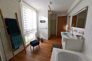 A bathroom with a bathtub, sink, and mirror. There is a window with blinds, a towel rack, and a wooden floor. - 45 to 90 m² gîte studio duplex, old-world charm dating from 1680, close to DINAN (Taden)