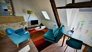 En modern lägenhet i loftstil med högt i tak, synliga träbjälkar, ett stort takfönster och en mysig sittgrupp med blå soffor och ett litet runt bord.