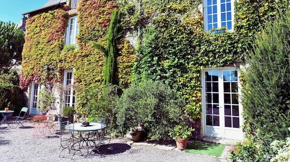 A building with ivy-covered walls, a gravel patio with a table and chairs, and a door with glass panels.