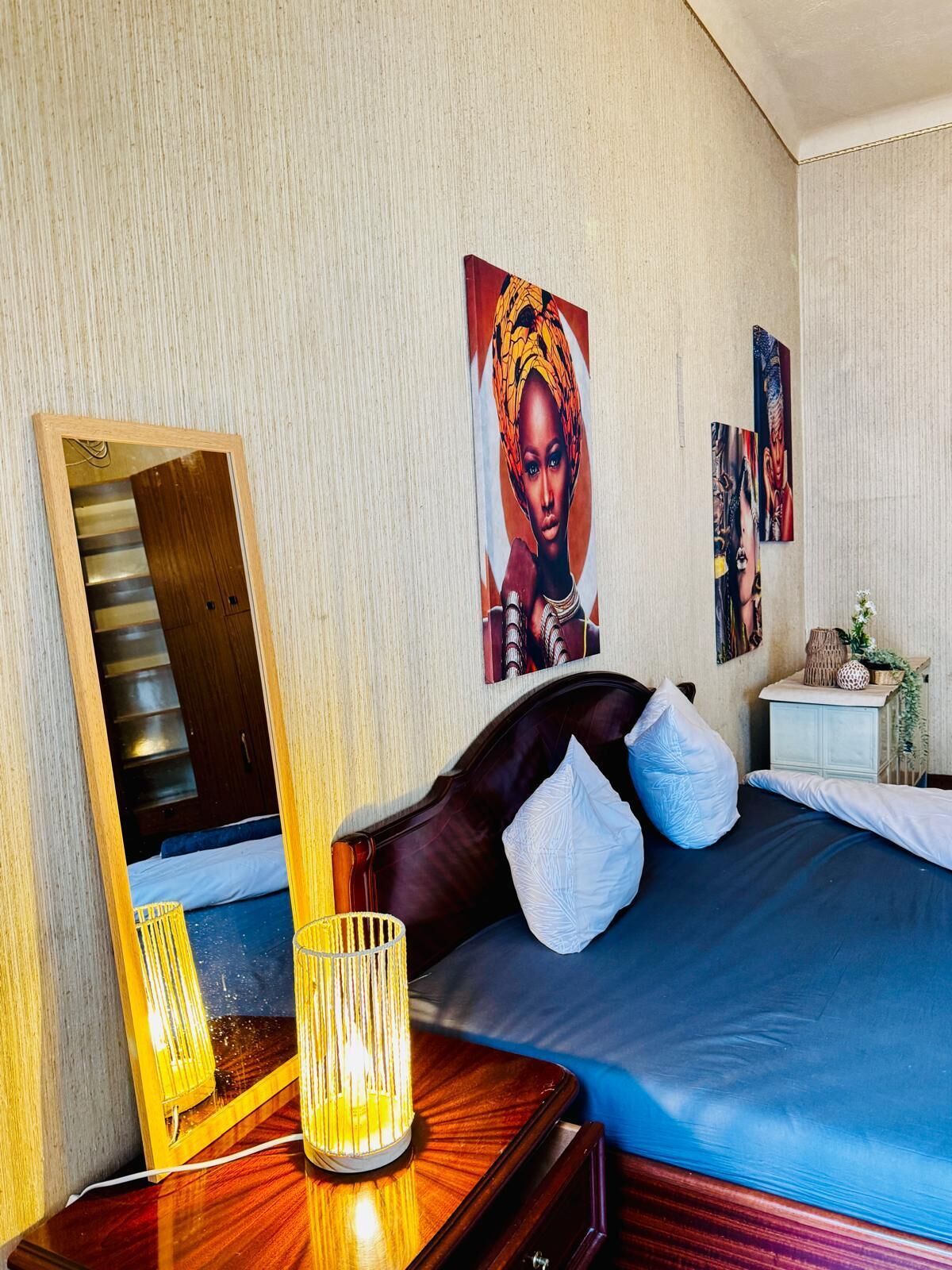 Quarto de hotel com uma cama grande, mesa de cabeceira com abajur, espelho e obras de arte nas paredes.