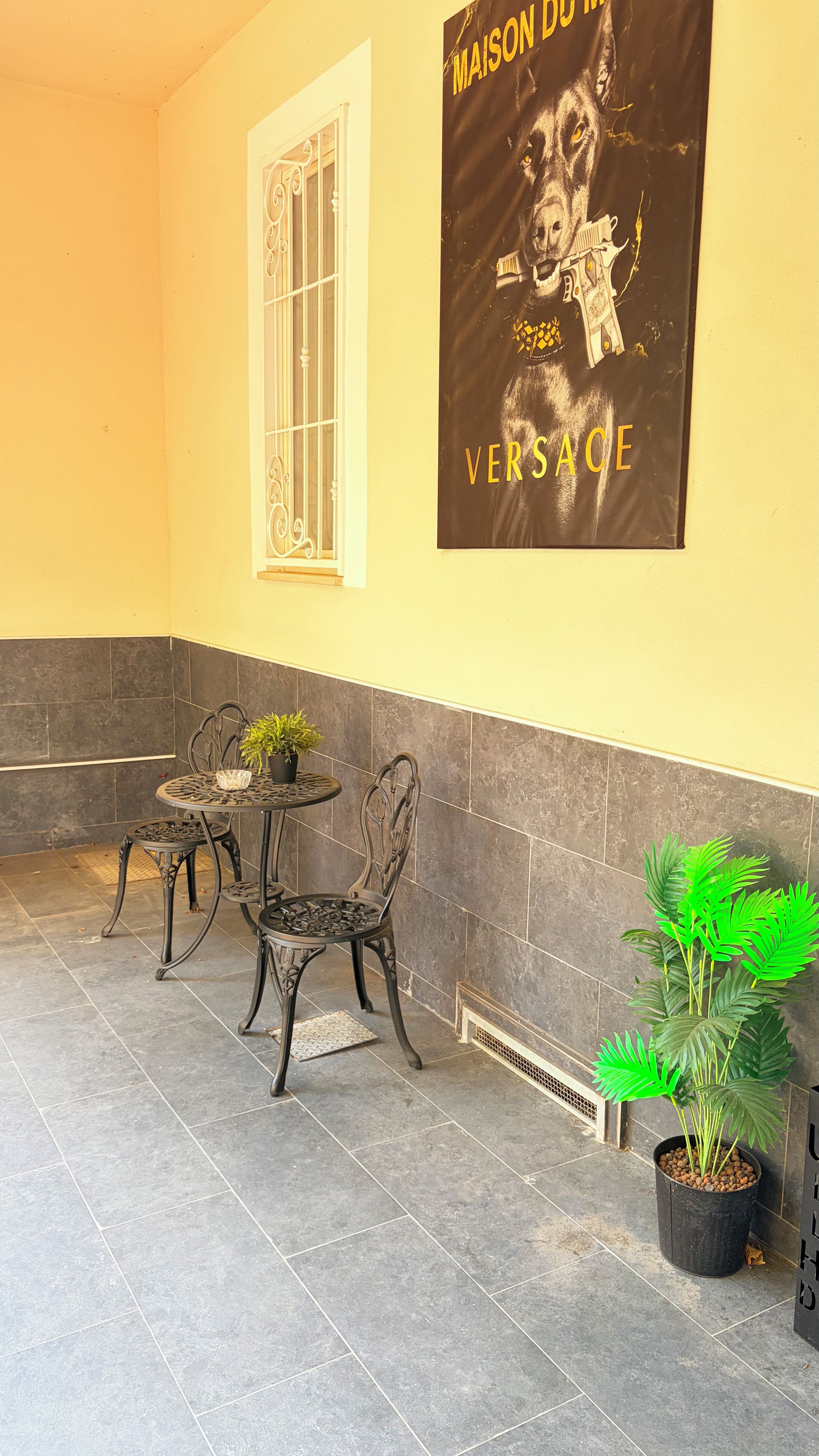 Een kamer met een betegelde vloer, een raam met witte kozijnen, een poster aan de muur, twee tafels met stoelen en potplanten.