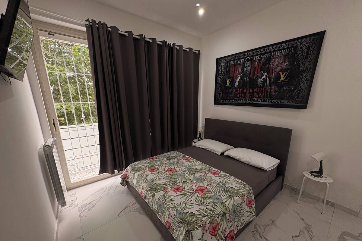 A bedroom with a bed, a window with curtains, a wall-mounted poster, and a small bedside table.