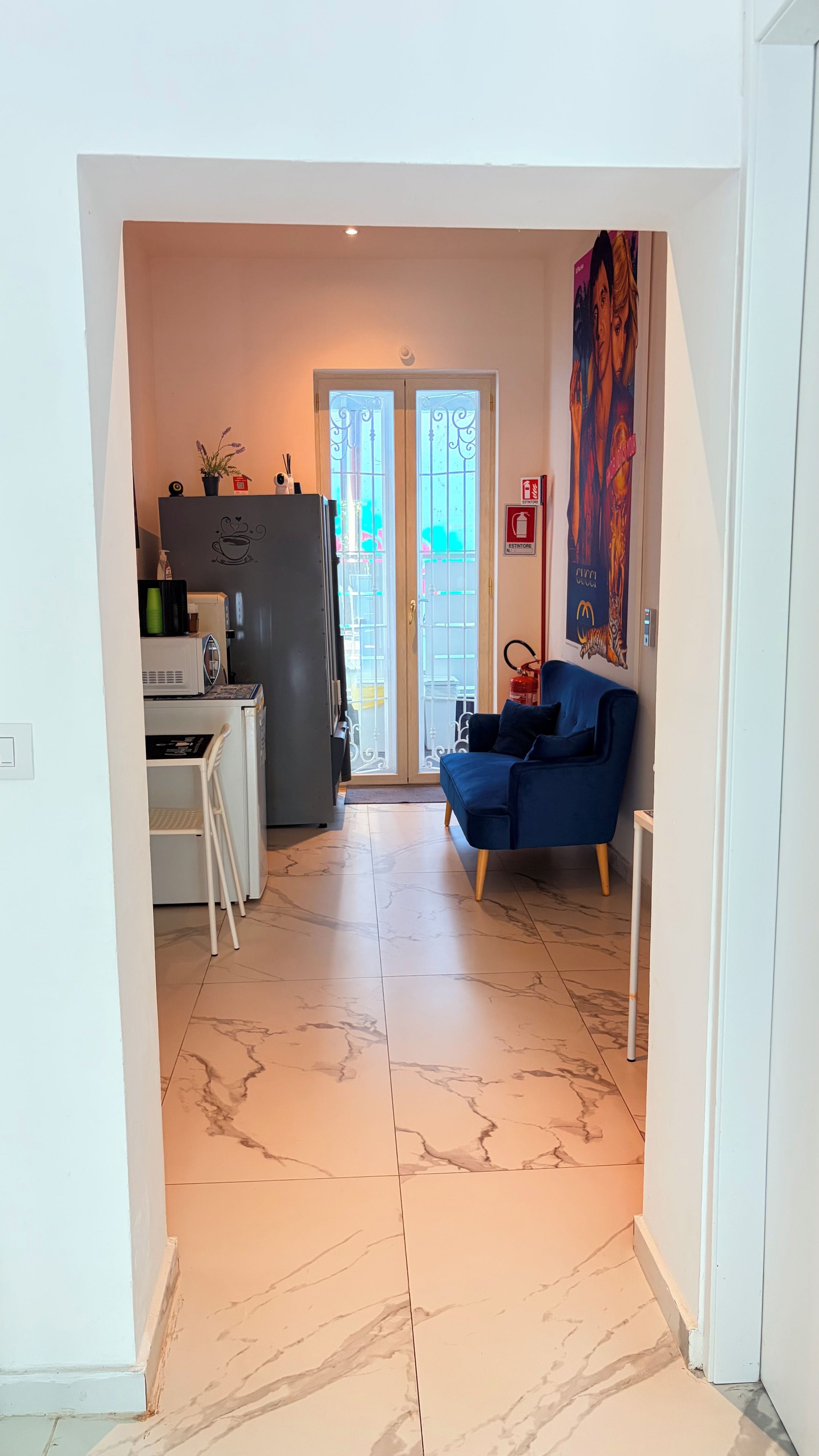 Una sala de estar con un sofá azul, un área de cocina con refrigerador y una puerta que da a un balcón.
