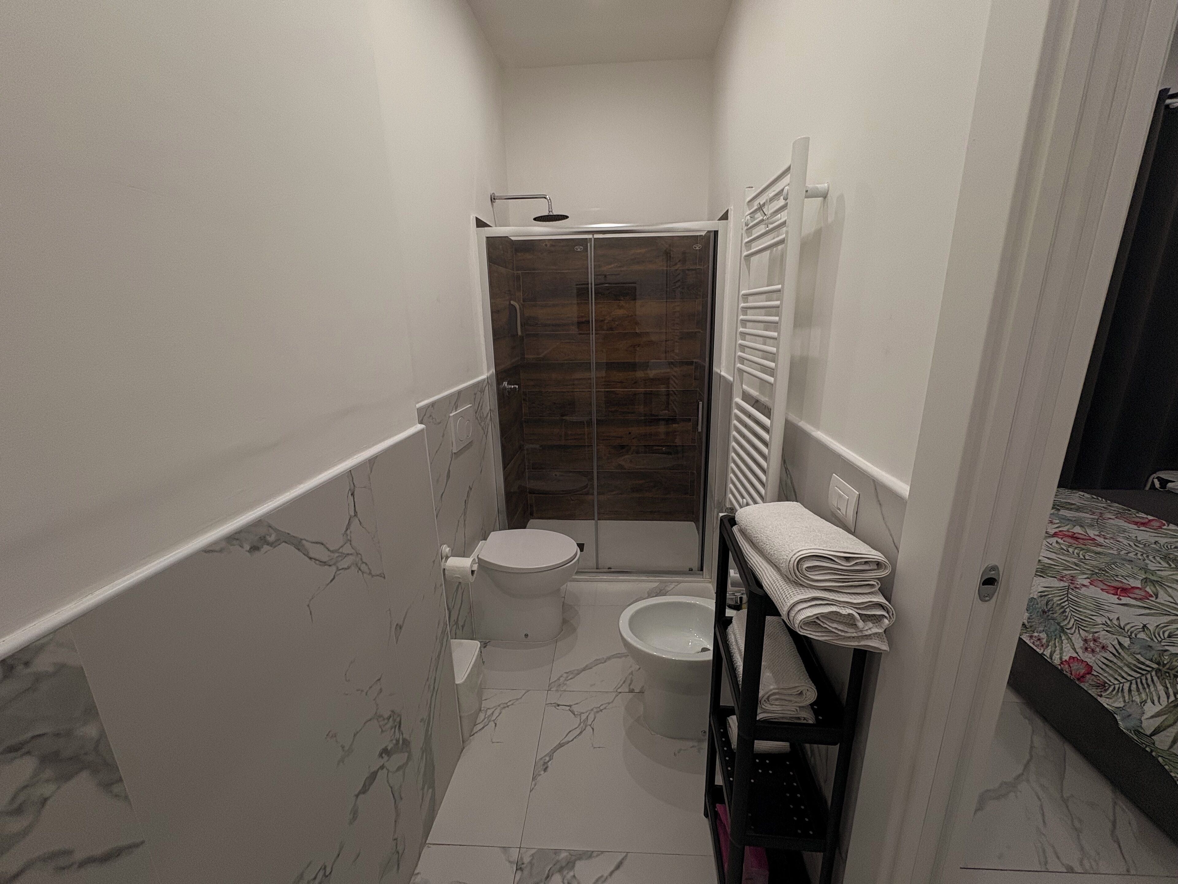 Ein schmales Badezimmer mit Toilette, Duschkabine und einem kleinen Ablagefach mit Handtüchern.