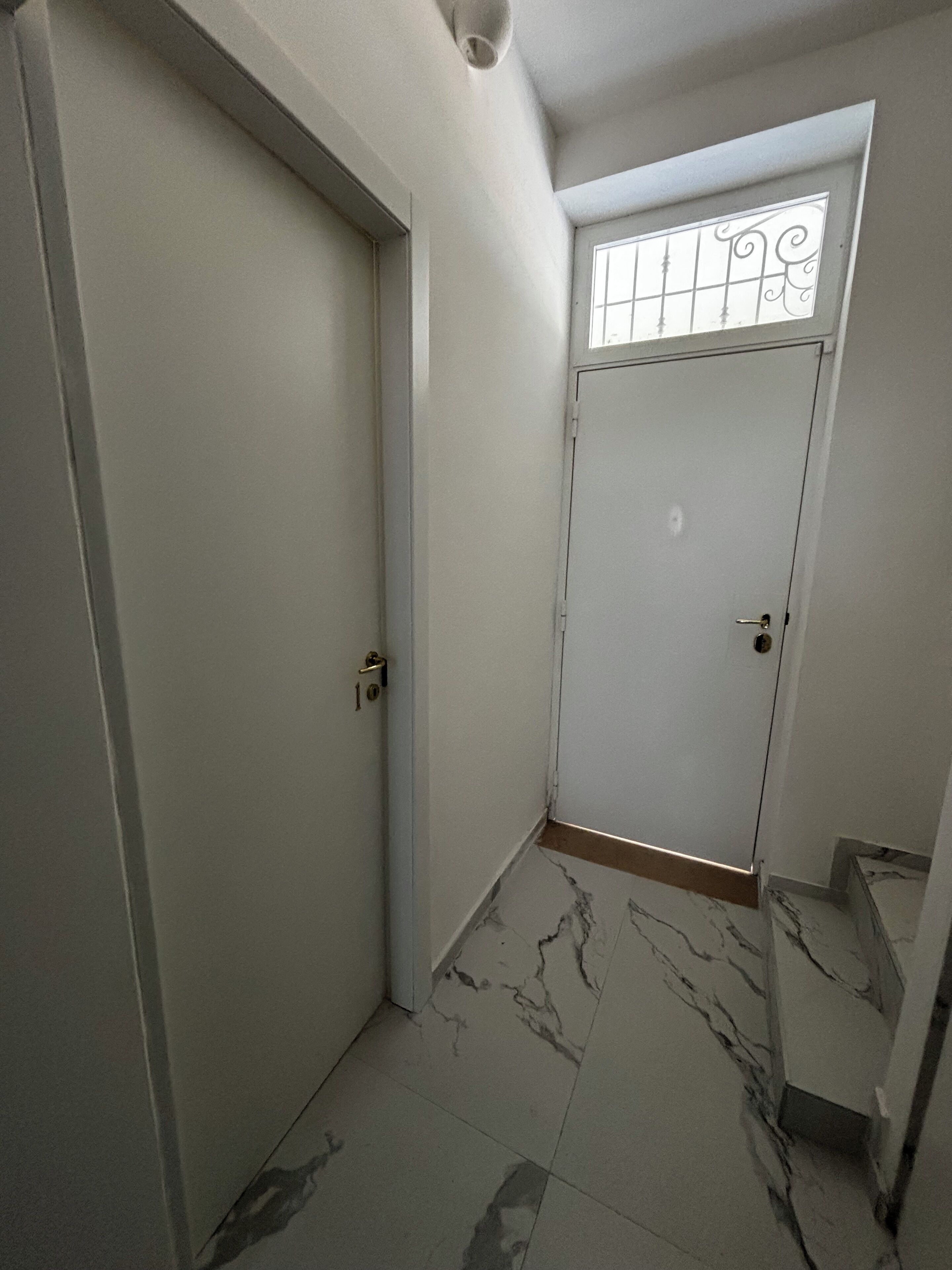 Un corridoio con una porta bianca, una finestra con inferriate decorative e un pavimento in marmo.