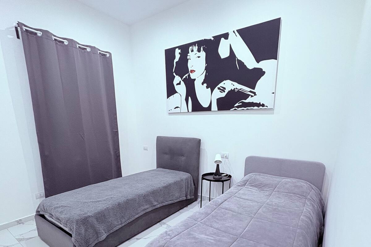 A modern bedroom with two beds, a grey curtain, a small table with a lamp, and a large black and white artwork on the wall.