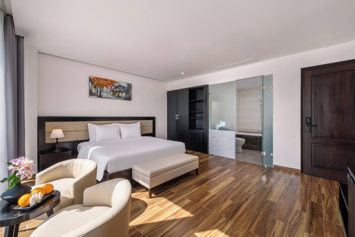 Une chambre d’hôtel moderne dotée d’un grand lit, d’un coin salon avec un fauteuil et une table, d’un petit balcon et d’une salle de bain avec douche et toilettes.