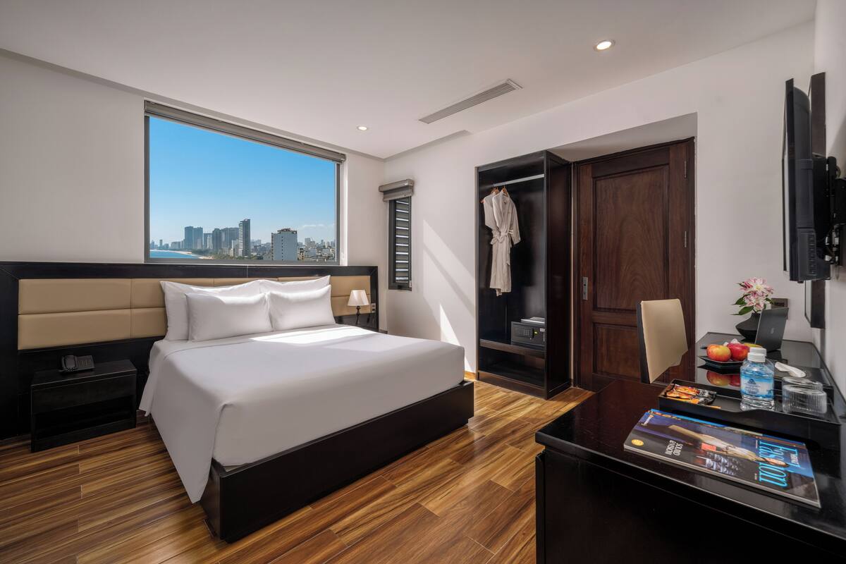 Une chambre d’hôtel moderne avec un grand lit, un bureau avec un magazine, un ordinateur portable et une vue sur le panorama de la ville.