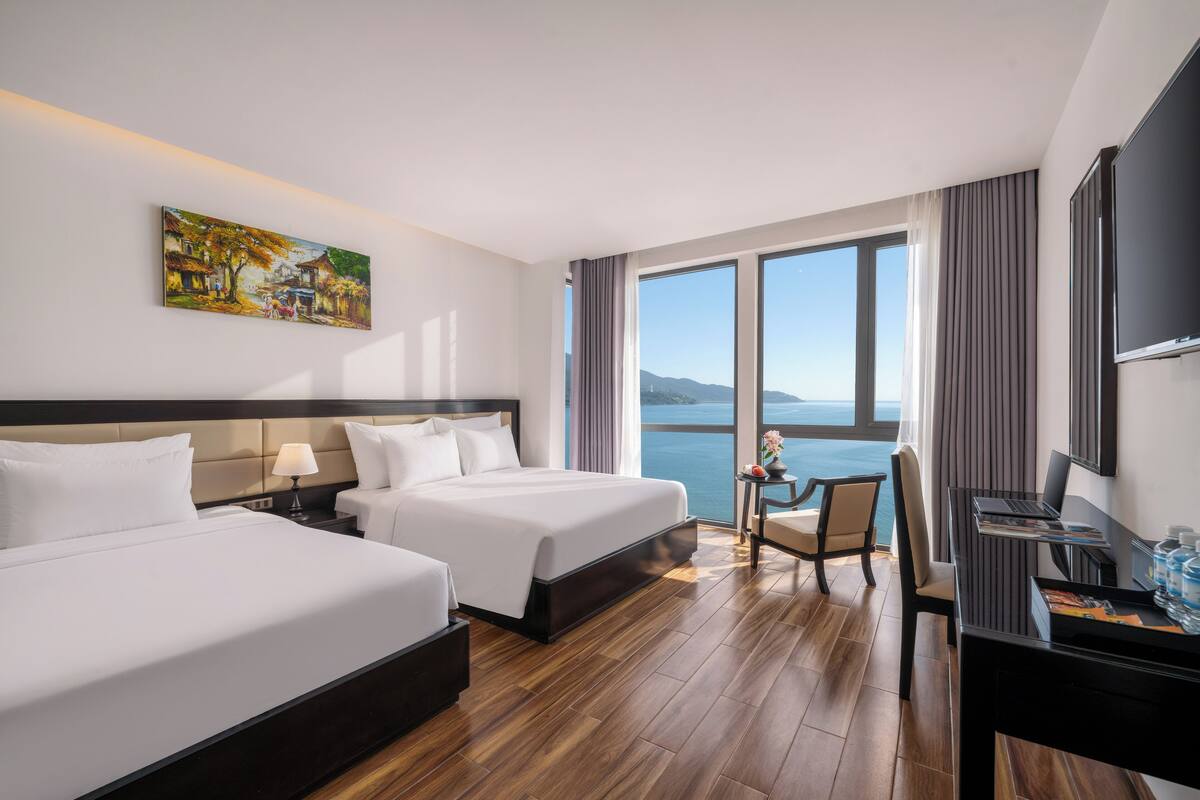 Une chambre d’hôtel avec deux lits, un bureau, une chaise, une télévision et une grande fenêtre donnant sur la mer.
