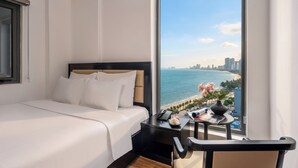 Une chambre d’hôtel avec un grand lit, une table de chevet, une chaise, une petite table sur laquelle se trouvent un vase et un magazine, et une vue sur l’océan et le paysage urbain.