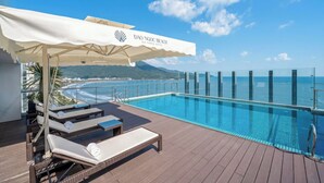 Un espace piscine aménagé sur le toit, avec des chaises longues et un auvent, offrant une vue sur une plage et l’océan.