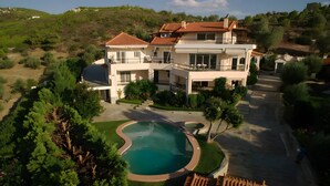 Une grande maison à deux étages avec une piscine, entourée de verdure et de collines.