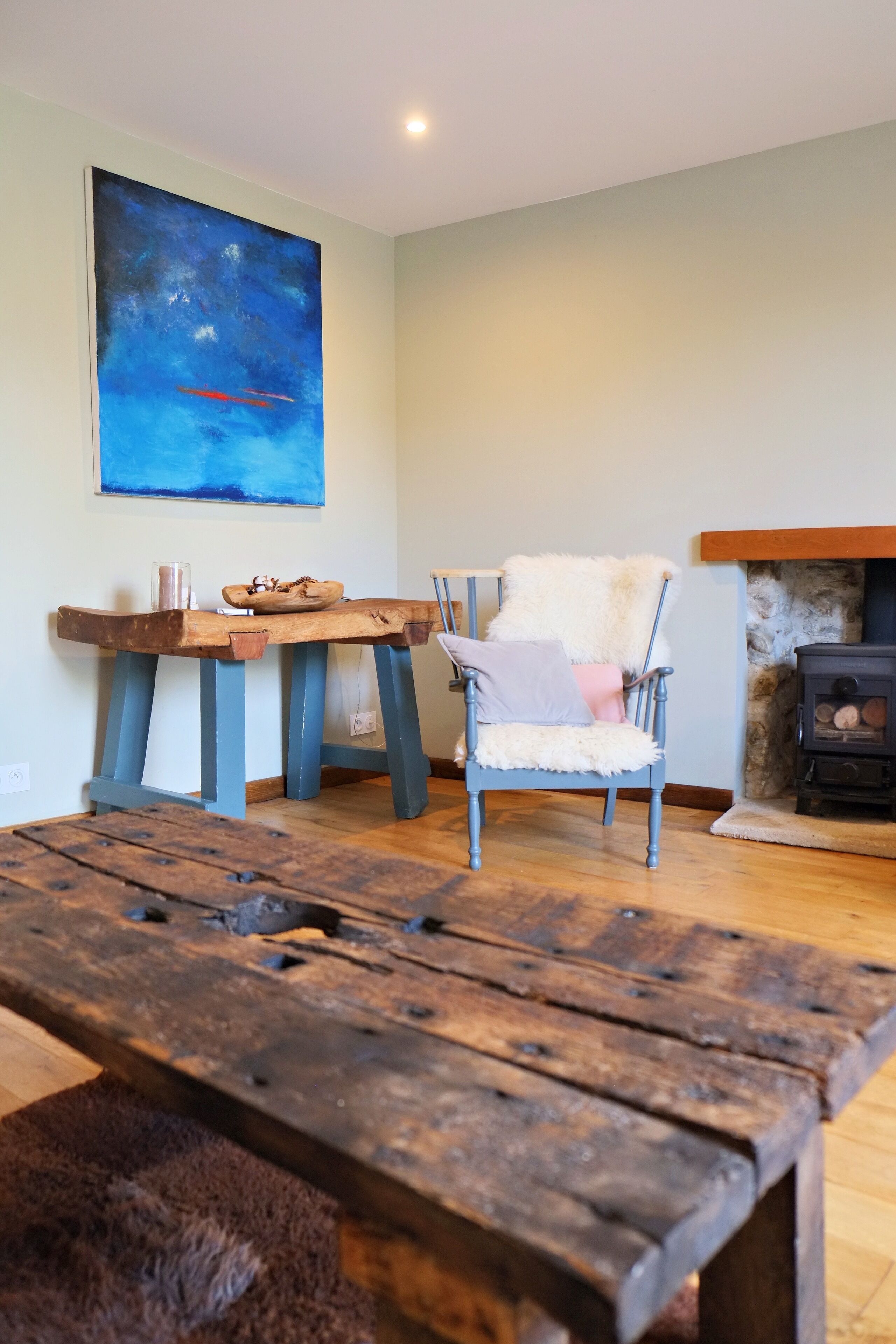 A rustic interior with a wooden table, a fluffy chair, a painting, and a stone fireplace.