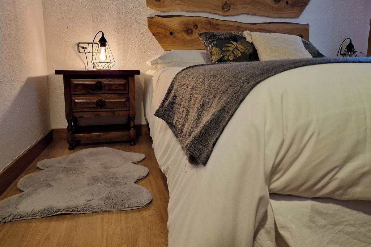 A bedroom with a wooden headboard, a bedside table with a lamp, a fluffy rug, and a bed with white bedding.