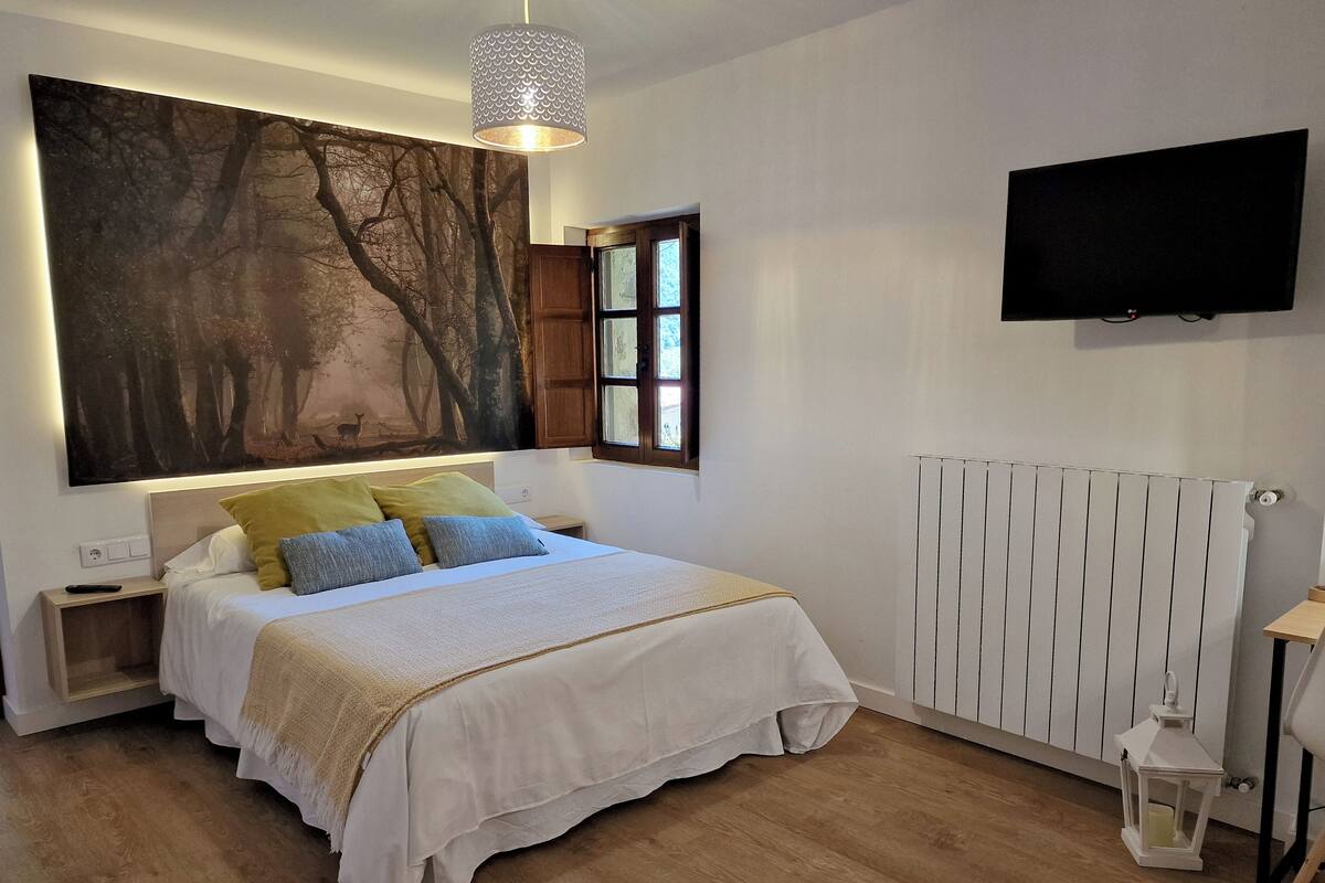 A bedroom with a large bed, a painting of trees, a window, a radiator, and a TV on the wall.