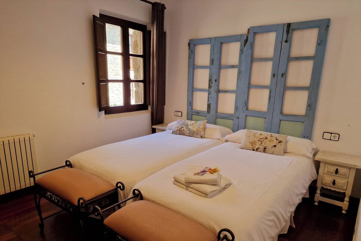 A double bed with a floral pillow, a bedside table, a radiator, and a window with curtains.