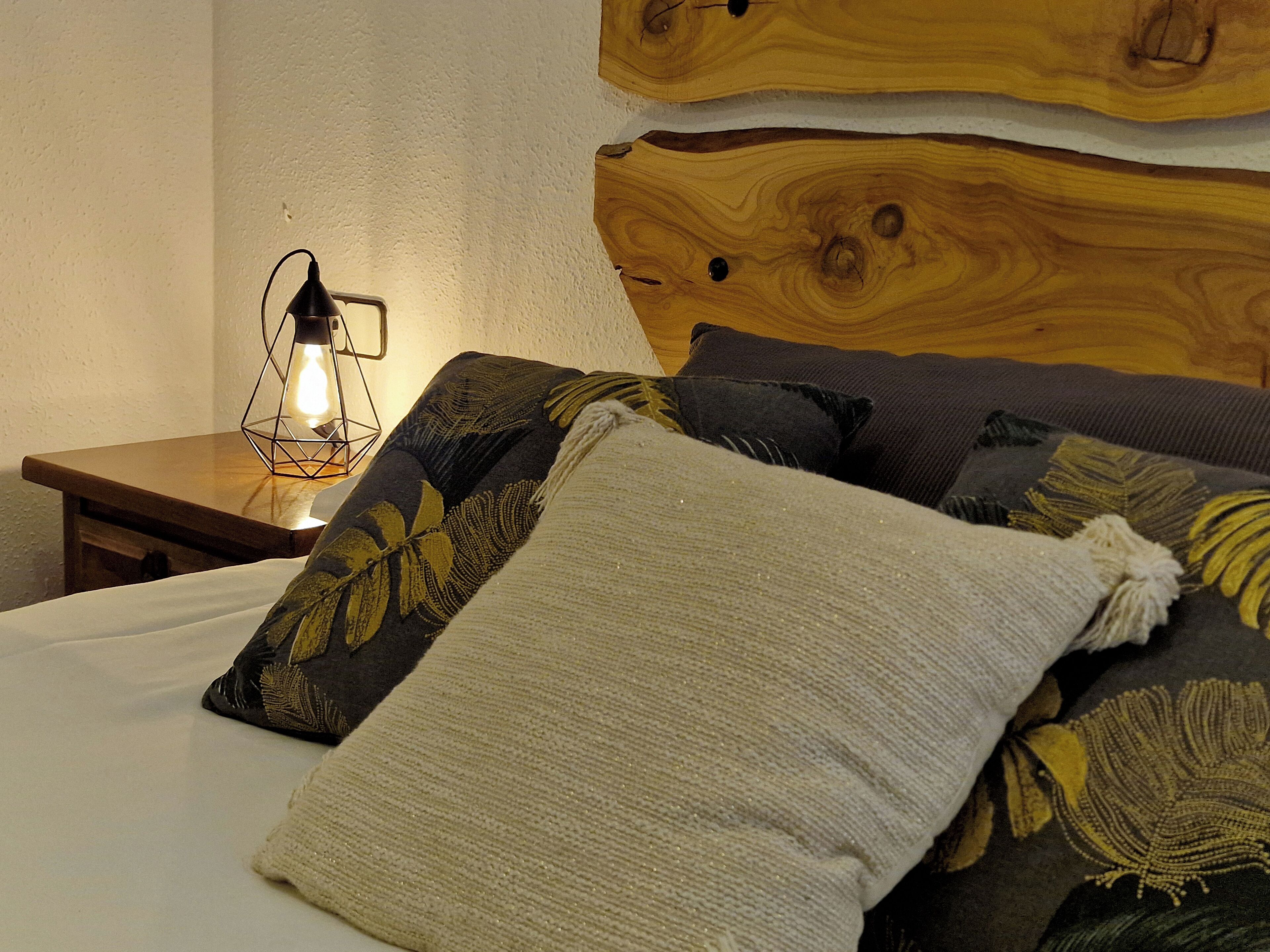 Una cama con cabecera de madera, una mesita de noche con lámpara y almohadas estampadas.