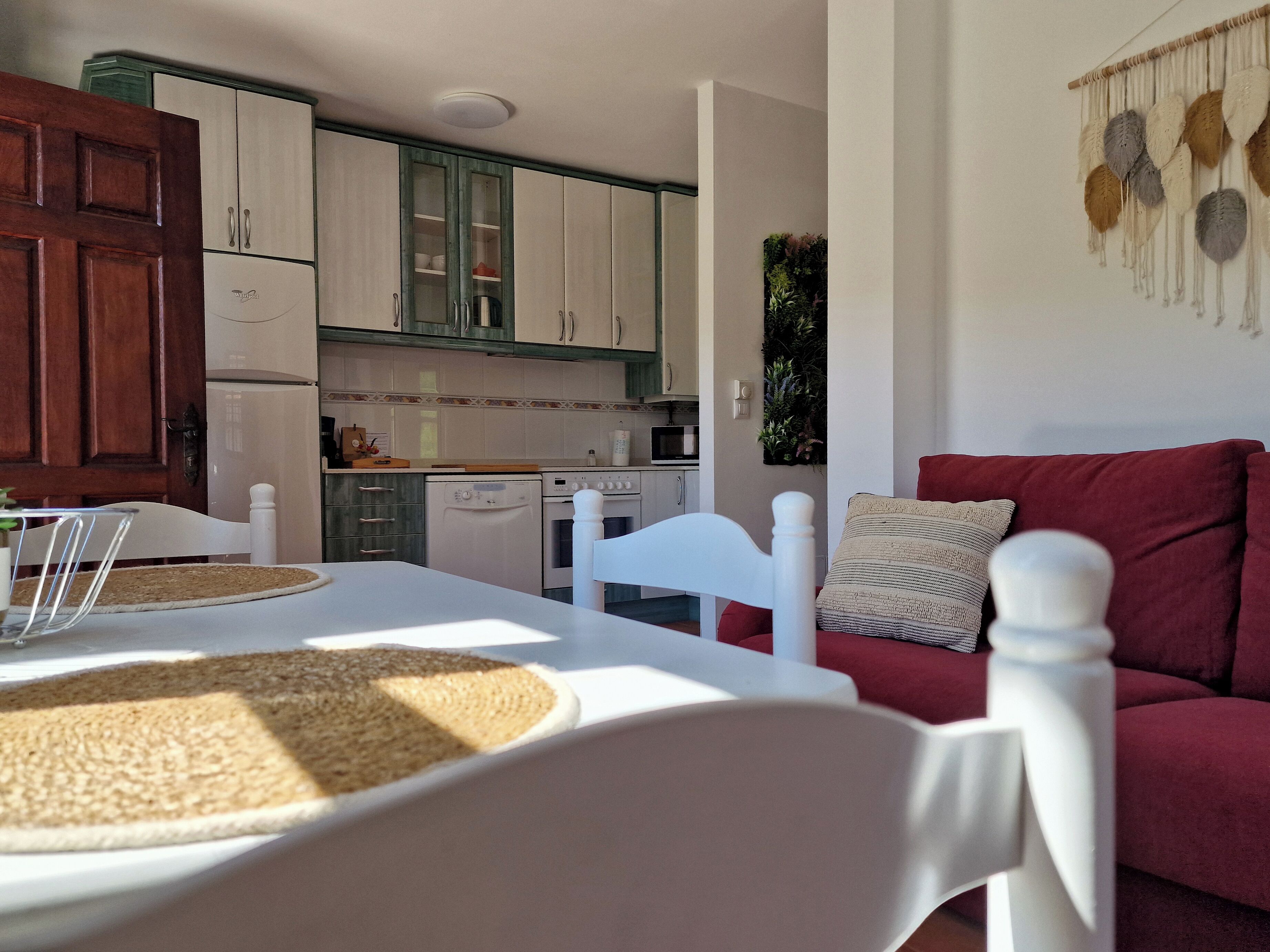 Une cuisine compacte avec des meubles blancs, un four à micro-ondes et un lave-vaisselle. On y trouve une table à manger avec une chaise blanche et un canapé rouge dans l’espace salon.