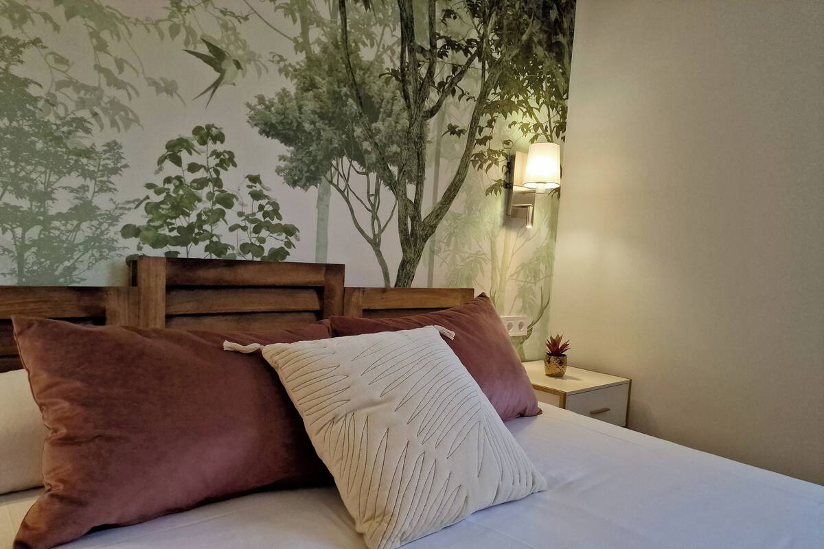 A bed with a wooden headboard, two pillows, a nightstand with a lamp, and a wall with a tree mural.