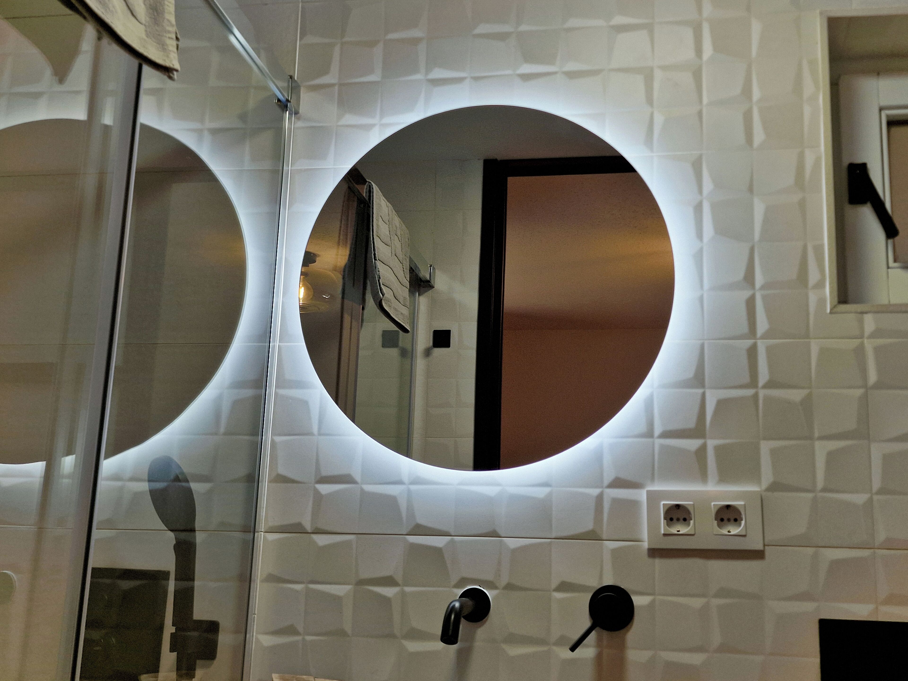 A bathroom with a circular mirror, a glass shower enclosure, and a white tiled wall with a geometric pattern.