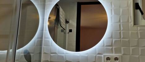 A bathroom with a circular mirror, a glass shower enclosure, and a white tiled wall with a geometric pattern.